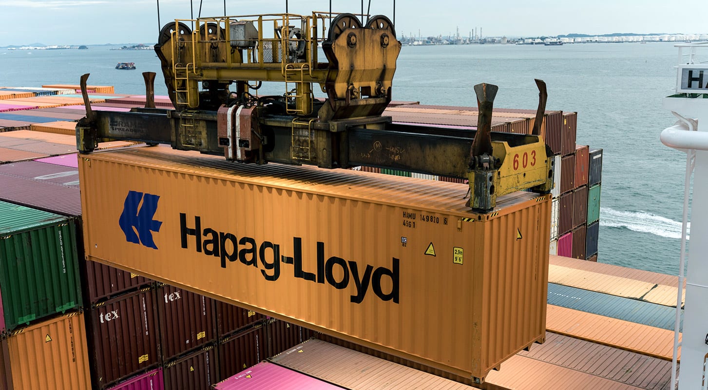 A large container ship is fully loaded with colorful Hapag-Lloyd shipping containers stacked on its deck.